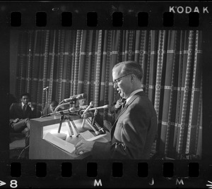 A man seen in profile, with his notes visible, speaking from a podium