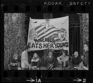 Student protesters sitting under an 'America Eats Her Young' banner