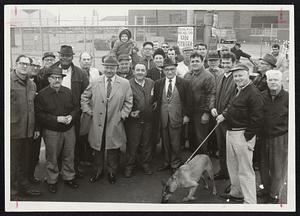 Dog on Leash pays visit to pickets patrolling outside the General Electric Company’s Lynn Riverworks plant in strike now in its fourth week. In center behind dog is Salvatore Camelio, president of the Massachusetts State Labor Council, AFL-CIO.
