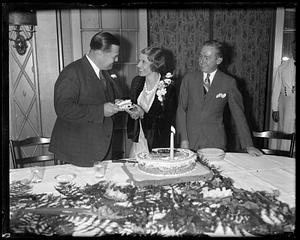 A man hands a woman a piece of cake as another man looks on
