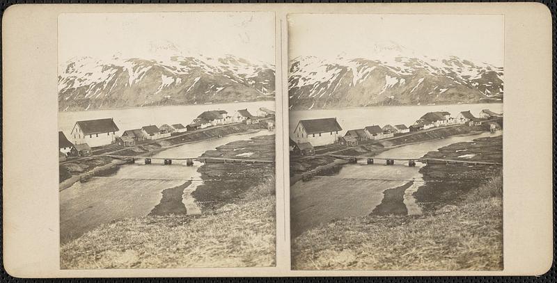 A group of houses on land surrounded by water, mountains in the background, a bridge in the foreground