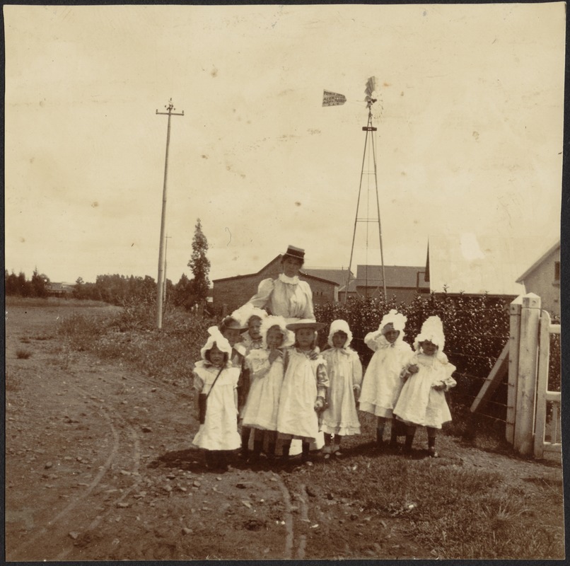 Woman with several young girls in white frocks standing in street near windmill