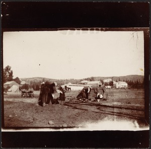 Group of people at camp site; wheel barrow and farm buildings in distance