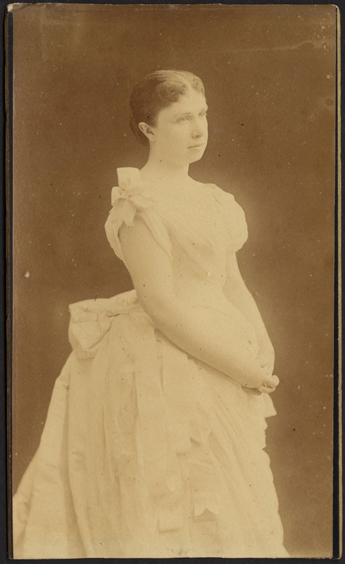 Young woman in white dress standing with hands folded; high bustle