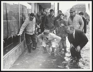Gov. John Volpe, center, bends down to look into a flooded store in Weymouth Landing. At left, Selectman William Rennie shows what the high water mark was at the height of the flood. Much of the area still is inundated. Gov. Volpe donned boots to tour the stricken areas.