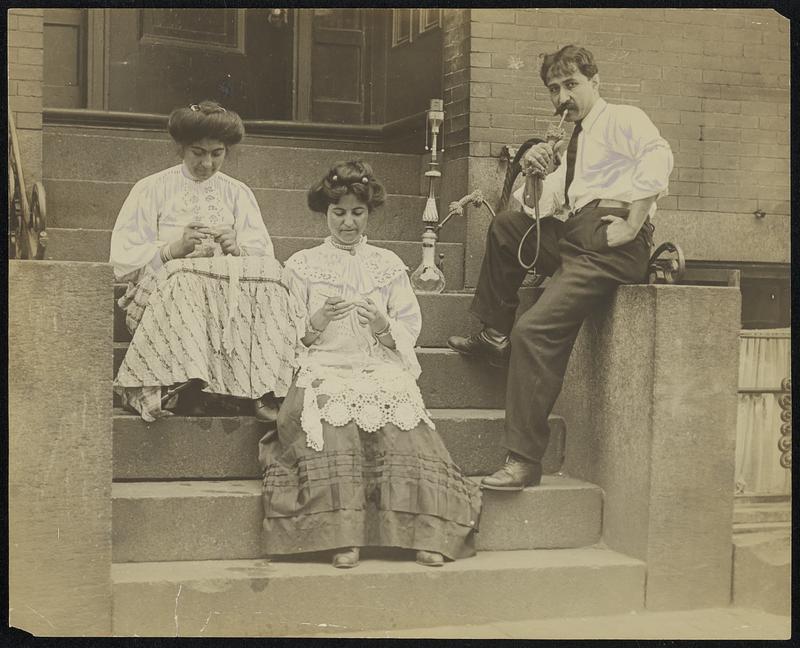 Three Turkish immigrants relax on the stoop of a tenement building. The two female figures are seated, their eyes on needlework in their hands. The third figure, a man, looks directly at the camera while smoking.