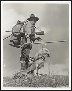 The Lure of Yukon Gold. Loaded with bedding, grub, pick, shovel, rifle, and gold pan, this old klondiker, accompanied by his pack-doc, prospects through the hills along the Yukon River, near forty mile, Canada, in his search for gold.