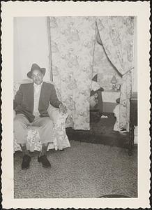 A man sits in a floral armchair by curtain