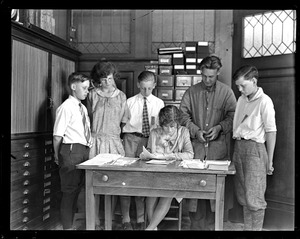 Child sits at desk reading papers while children stand behind her and observe