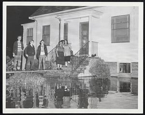 Dedham Folks Smiling Through although marooned by flood. Mrs. Anna Tuzzo, center, stands on steps of her Bridge street home with neighbors after Charles River had flooded her home. Boys, left to right, Charles Noff, Jerry Lowell and Walter Noff. With Mrs. Tuzzo are Mrs. Joan-McArdle and Mrs. Kerri George.