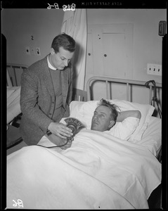 Unidentified man holding WAAB microphone in front of an unidentified man lying in a hospital bed