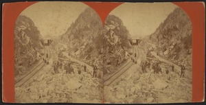 Train tracks cutting through two mountains, with rocks and boulders in foreground and people in midground