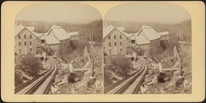View from mountaintop, looking down railroad tracks to a train at the platform and a cluster of buildings