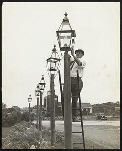 From Fred Roche, Cape Playhouse, Dennis, Mass. Last of the Lamplighters-- Charles Foley, carrying his little ladder and plenty of matches walks around the 28-acres estate of the Cape Playhouse, and Cape Cinema conducted by Raymond Moore at Dennis, Mass, twice nightly, one to light and once to put out the old fashioned kerosene lamps in these antique lamp posts. He is probably the last of a once numerous body of lamp-lighters whose daily task was to light the way for late travelers. The Cape Playhouse is seen in the background.