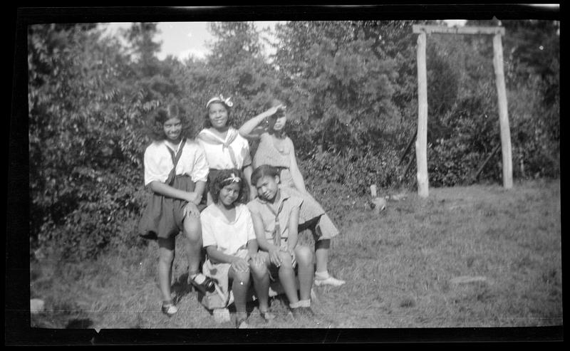 Five young women pose in front of foliage, wooden structure in background