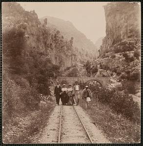 Sightseers walking along railroad tracks with bridge and mountains in background