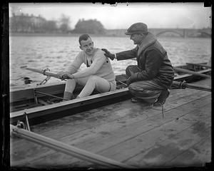 A member of Harvard crew sits in a shell on the Charles, Coach Charles Whiteside kneeling next to him, Larz Anderson Bridge and Weld Boathouse in background