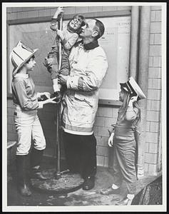 Children at a fire station, one being held on the fireman's pole by a fireman
