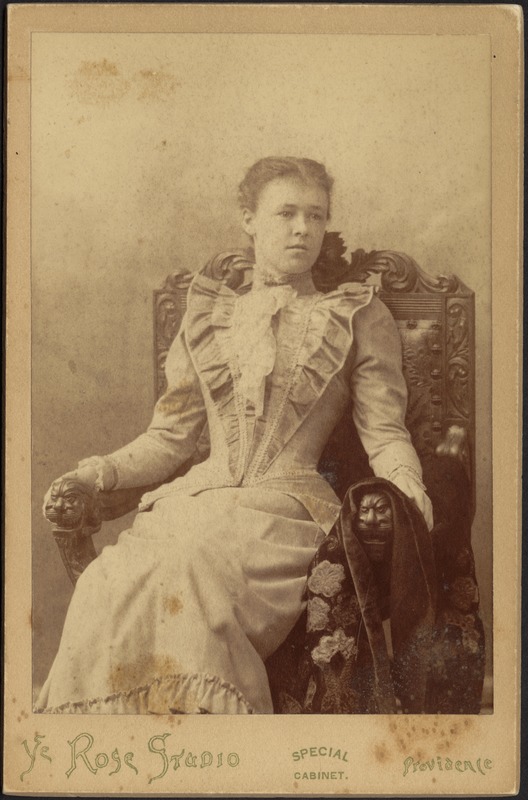 Young woman in light dress with white ruffle collar; seated in ornately carved wooden chair