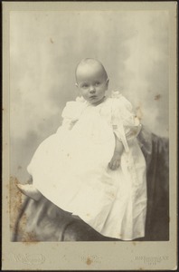 Baby in long white dress and white shoes seated on draped background