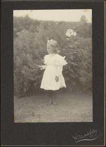 Young blonde girl in white dress and bow in hair, standing in garden