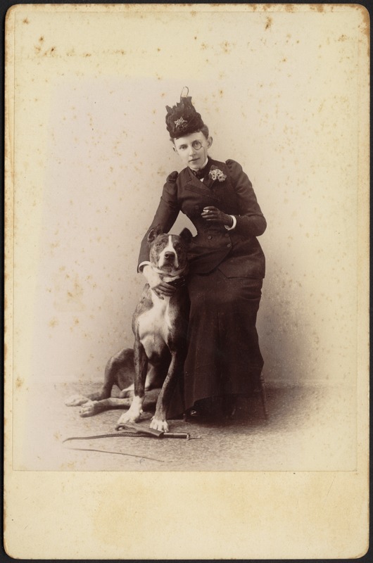 Studio portrait of unidentified woman in black dress and monocle with cigarette posing with Great Dane; whip and glove on floor