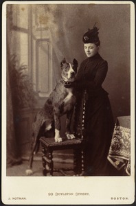 Studio portrait of woman in black dress standing near Great Dane on chair