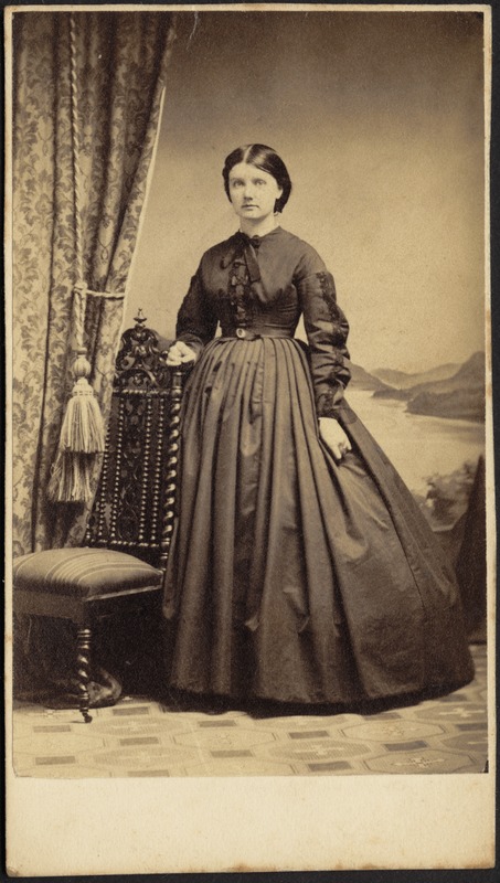 Portrait of woman in dark dress standing near chair and heavy drape