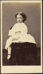 Young girl in white frock seated on dark chair with fringe