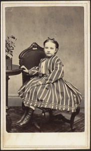 Young girl in striped dress seated in chair, hand on table