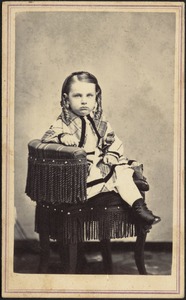 Young child wearing plaid frock seated in dark chair with fringe