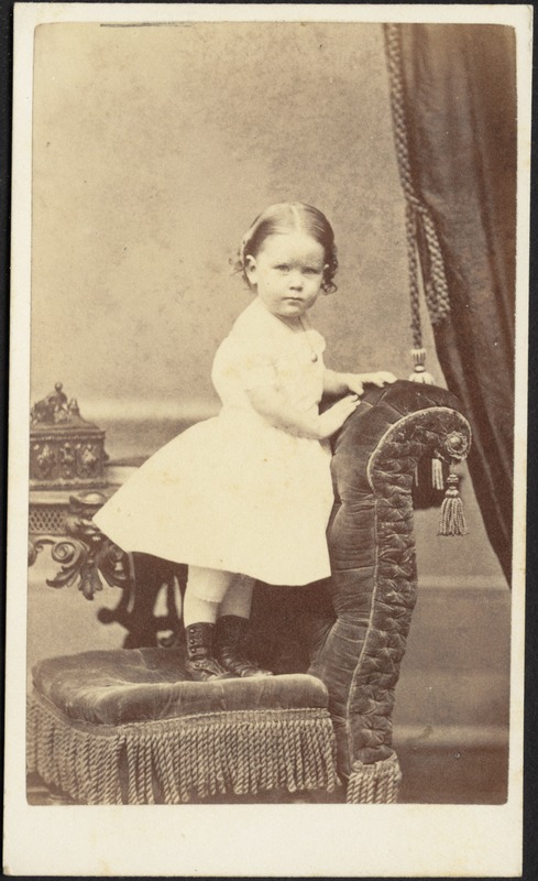 Young child in white frock, standing on upholstered chair with tassels
