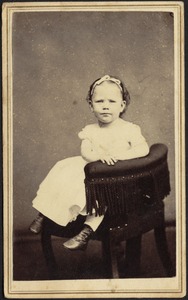 Young child in white frock seated in chair with fringe