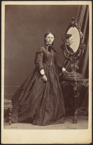 Woman in dark dress standing, her profile reflected in mirror
