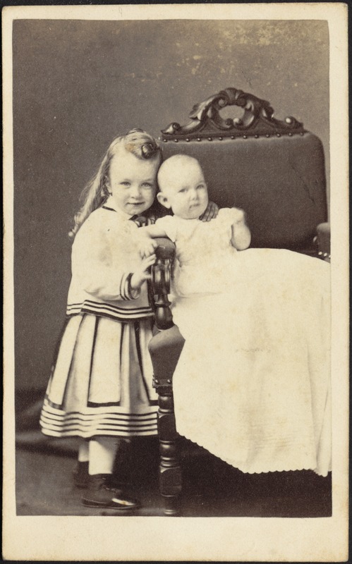 Child with long blond ringlets standing next to infant seated in high-back chair