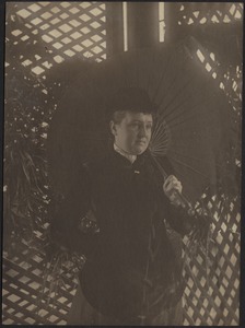 Woman in dark dress and hat holding parasol, wall of wooden lattice behind her
