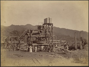 Four people (1 woman, 3 men—two with pith helmets) standing in front of a large wooden rig with stairs, belts, pulleys, pipes and tanks or engines with smokestacks (possibly a mill or pump, perhaps a method for cleaning ore or gold); mountains in distance