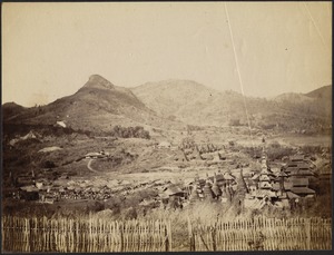View from wattle fence of rural Burmese village, featuring ancient stone temples with conical spires and a wooden pagoda structure; people and livestock gathered near straw huts on left; mountains in distance