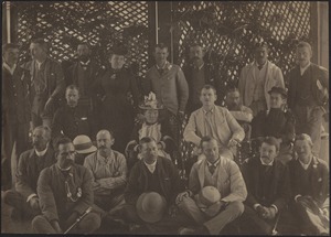 Group of twenty people (three women) posing in front of wooden lattice wall; some men with pith helmets