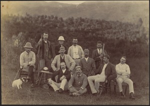 Group of eleven men seated outdoors; many with mustaches and some with pith helmets; small white dog on left