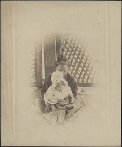 Asian woman holding Caucasian infant in white bonnet in lap, wooden lattice wall