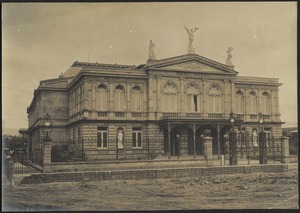 View of façade of "Teatro Nacional de Costa Rica" from dirt road; brick retaining wall in front of cast iron fence (opened in 1897)