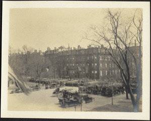 View looking across of city plaza filled with cars and people; street lined with brownstones; American flag on left in foreground