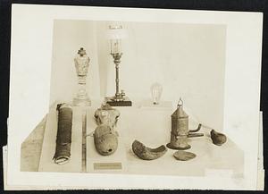 Many Instruments Have Aided Man in Securing Light. The Smithsonian Institution has recently arranged an exhibit showing the evolution of light from the days of the campfire to the present. Specimens in the collection show many weird forms of lamps and torches in vague at different periods. The torch at the left of the photograph is of birch bark, while next to it are a gourd and the skull of an animal. Later stones were fashioned into peculiar shapes to hold grease and oil. Oil was later burned in tin vessels followed by wicks inserted in glass receptacles. The latest development is that of the modern incandescent bulb and the modern gas fixture.