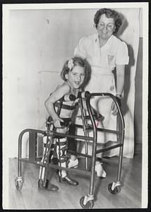 Polio Victim Stands Up-Janice Dawe, five-year-old English polio victim who was brought to this country in the hope of being cured, takes a few steps with the aid of Miss Dorothy Wagner, physio-therapist at Michael Reese Hospital in Chicago. Aided by new braces and a mobile support frame, the child stood up for the first time in two and a half years. Her parents sold their home and belongings to finance the trip.