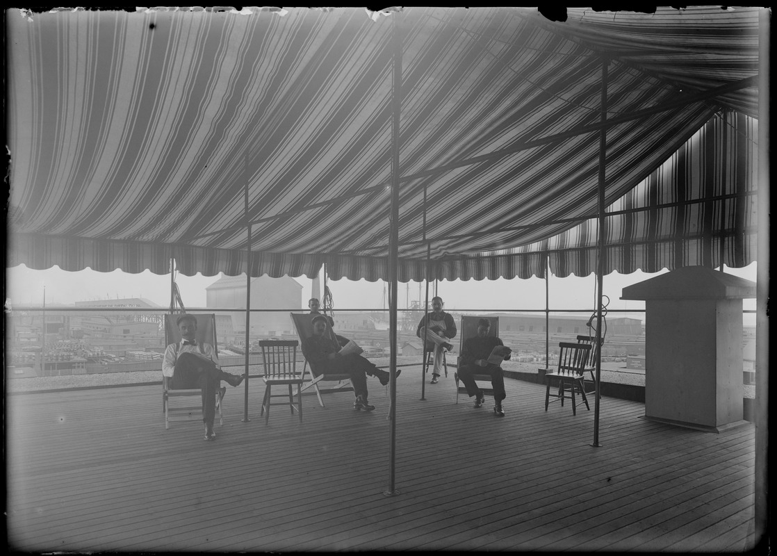 Group of men sitting under roof-top pavilion, wharves in background - grain elevator, ship, freight cars, (B.W.C. employees?)