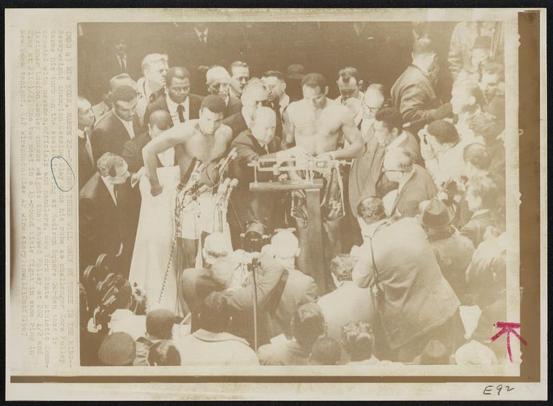 Tonight There Will Only By Three in the Ring--Heavyweight champion Cassius Clay dons his robe as challenger Zora Folley takes his turn on the scales in ring at Madison Square Garden that is crowded with newsmen, officials and handlers. New York State Athletic Commissioner Edwion Dooley checks weights that showed Folley at 202 1/2 and Clay at 211 1/2. The two meet in a 15-round title fight in same ring in New York tonight.