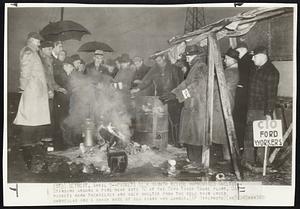 Detroit – Pickets Seek Warmth Beside Improvised Shelter – Standing around a fire near gate 12 at the Ford River Rouge plant, UAW pickets warm themselves and seek shelter from the cold rain under umbrellas and a shack made of old signs and lumber.