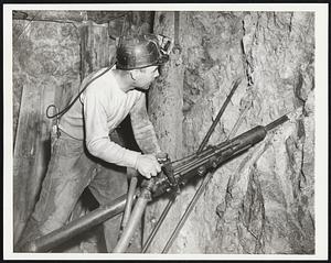 A Vital Wartime Metal - This industrial soldier is a hard rock miner producing manganese ore for use in wartime steel production. His "gun" is a compressed air-operated drill with a water connection to eliminate dust. He is drilling in the face of the drift at the Emma mine, operated here by the Anaconda Copper Mining Company.