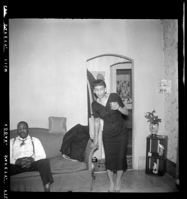 Woman stands holding a glass next to a man in suspenders seated on a sofa